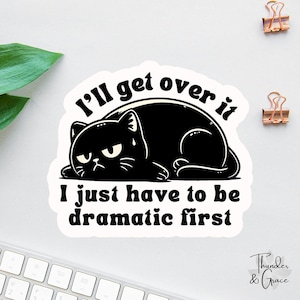May include: A white sticker with a black cat illustration. The cat has an angry expression and is lying down. The text on the sticker reads "I'll get over it, I just have to be dramatic first."