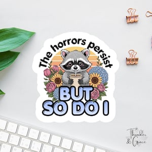 Pode incluir: Um autocolante com um guaxinim de desenho animado bebendo de uma chávena, rodeado por flores. O texto diz "The horrors persist BUT SO DO I". O design inclui um fundo de pôr do sol e está rotulado "Thunder & Grace".