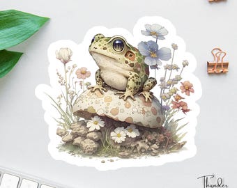 Könnte beinhalten: Ein grüner und brauner Frosch sitzt auf einem Pilz mit weißen Flecken. Der Frosch hat große, schwarze Augen und ist von zarten, pastellfarbenen Blumen umgeben.