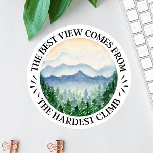 Op de afbeelding: Een ronde witte sticker met een aquarel-illustratie van een bergketen en een bos. De tekst "THE BEST VIEW COMES FROM THE HARDEST CLIMB" staat in het zwart rond de afbeelding.