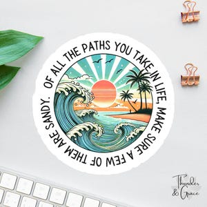 May include: Round sticker with a beach scene featuring a sunset, ocean waves, and palm trees. The sticker has the text "Of all the paths you take in life, make sure a few of them are sandy."