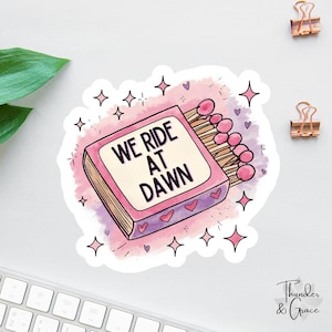 May include: A sticker featuring a pink matchbox with the text "WE RIDE AT DAWN" in black. The matchbox is surrounded by pink stars and hearts. The matches are pink and brown.