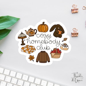 May include: A die-cut sticker featuring the text "cozy homebody club" surrounded by illustrations of a pumpkin, a brown rabbit, a lamp, a plaid blanket, a brown sweater, a basket of red apples, a cinnamon roll, and autumn leaves.