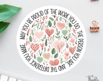 May include: A round sticker with a floral and heart design in pink, green, and white. The sticker has the text "May you be proud of the work you do, the person you are, and the difference you make." Two gold binder clips are in the background.