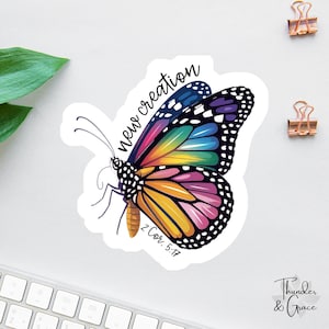 May include: A colorful butterfly sticker with a rainbow wing design and the words "new creation" in black script. The sticker also includes the Bible verse "2 Cor. 5:17". The sticker is on a white surface.