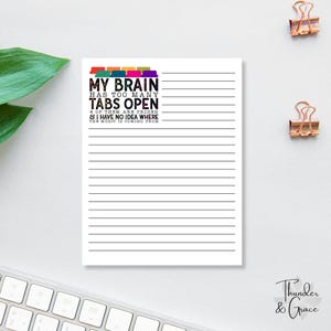 May include: A white lined notepad with the text "MY BRAIN HAS TOO MANY TABS OPEN" in black, with colorful tabs above the text. The notepad also has the text "4 OF THEM ARE FROZEN & I HAVE NO IDEA WHERE THE MUSIC IS COMING FROM". Two gold paper clips are in the background.