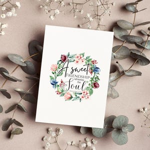 May include: A white greeting card with the text "A sweet friendship refreshes the Soul" surrounded by a floral wreath in pink, blue, and burgundy. The card is on a light pink surface with sprigs of greenery.