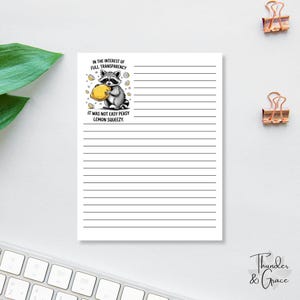 Op de afbeelding: Wit gelijnd notitieblok met een cartoon wasbeer die een citroen vasthoudt. Het notitieblok heeft de tekst "IN THE INTEREST OF FULL TRANSPARENCY" en "IT WAS NOT EASY PEASY LEMON SQUEEZY." Twee gouden paperclips zijn zichtbaar.