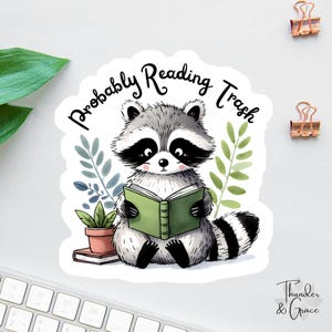 Könnte beinhalten: Ein Aufkleber mit einem Cartoon-Waschbären, der ein grünes Buch liest. Der Waschbär hat schwarzes und graues Fell und ist von grünen Blättern und einer kleinen Topfpflanze umgeben. Der Text "Probably Reading Trash" steht über dem Waschbären.