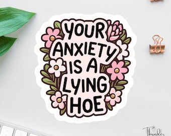 Può includere: Un adesivo con la scritta "YOUR ANXIETY IS A LYING HOE" in nero, circondata da fiori rosa e foglie verdi. L'adesivo ha un bordo bianco ed è su una superficie bianca. Sono visibili due fermagli dorati.