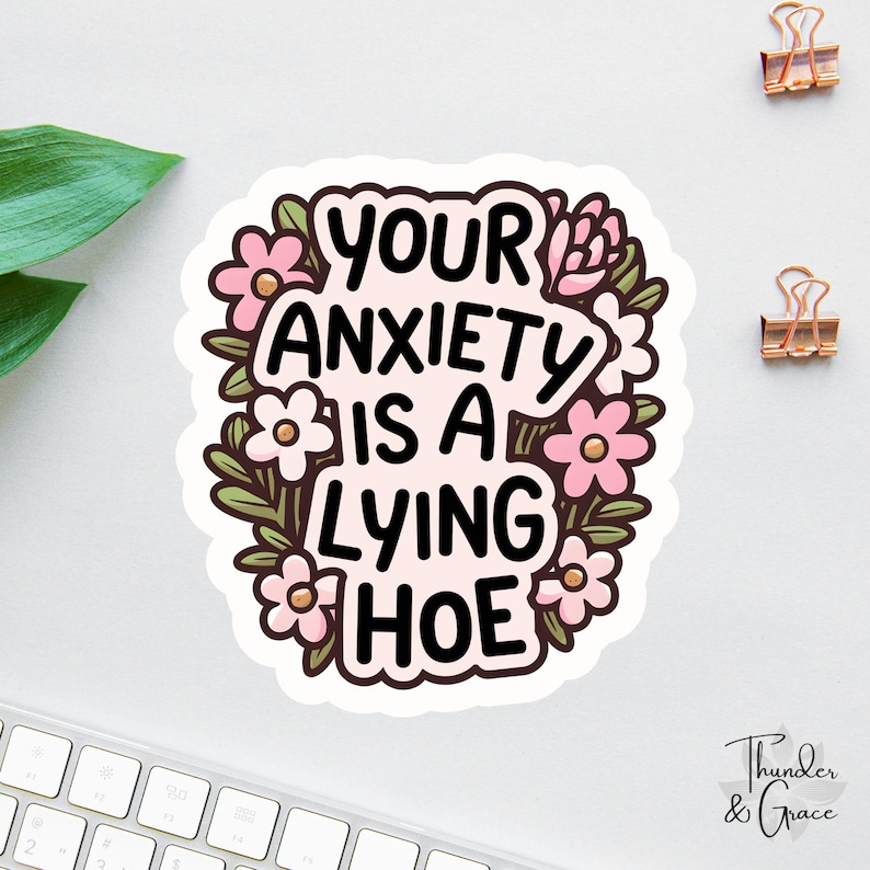 May include: A sticker with the text "YOUR ANXIETY IS A LYING HOE" in black, surrounded by pink flowers and green leaves. The sticker has a white border and is on a white surface. Two gold paper clips are visible.