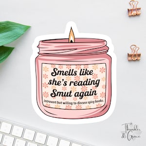 May include: A pink candle sticker with the text "Smells like she's reading Smut again." The sticker has a floral design and the phrase "introvert but willing to discuss spicy books." The candle has a lit wick.