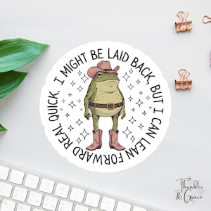 Peut inclure: Un autocollant circulaire représentant une grenouille verte portant un chapeau de cowboy marron, une ceinture marron et des bottes de cowboy roses. La grenouille est entourée de petites étoiles et du texte, I MIGHT BE LAID BACK, BUT I CAN LEAN FORWARD REAL QUICK.