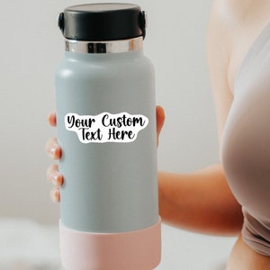 May include: A gray reusable water bottle with a pink silicone base. A white sticker with the text "Your Custom Text Here" is on the bottle.