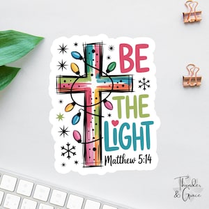May include: A colorful sticker featuring a cross decorated with string lights. The text "BE THE LIGHT" is displayed in various colors, with "Matthew 5:14" below. The design includes snowflakes and stars.