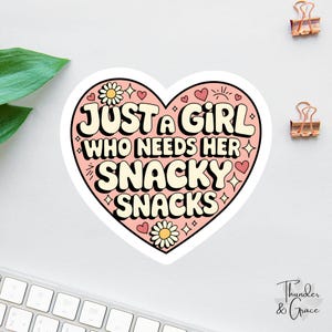 May include: A heart-shaped sticker with the text "JUST A GIRL WHO NEEDS HER SNACKY SNACKS" in a retro font. The sticker is pink with white and black accents, featuring daisies and hearts. Two gold paper clips are in the upper right corner.
