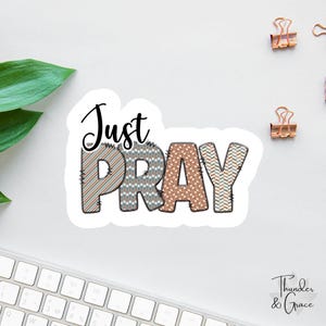 May include: A white sticker with the words "Just PRAY" in a mix of patterned fonts. The letters are in shades of brown, gray, and white. The sticker is on a white surface with a keyboard and paper clips in the background.