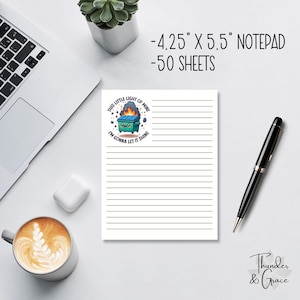 May include: A lined notepad with the text "THIS LITTLE LIGHT OF MINE I'M GONNA LET IT SHINE" and a cartoon dumpster graphic. The notepad measures 10.8 cm x 14 cm and has 50 sheets. A black pen and a cup of coffee are also visible.
