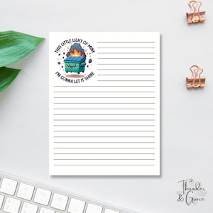 May include: A lined notepad with a cartoon illustration of a smiling dumpster with flames and the text "This little light of mine, I'm gonna let it shine." The notepad has a white background and black lines.