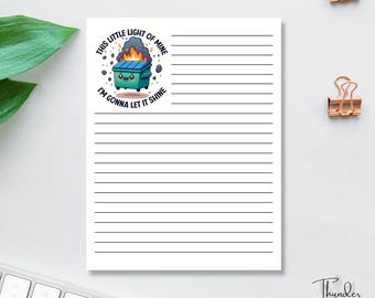 May include: A lined notepad with a cartoon illustration of a smiling dumpster with flames and the text "This little light of mine, I'm gonna let it shine." The notepad has a white background and black lines.