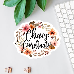 May include: A white sticker with a floral wreath design. The wreath is made of brown branches, orange and pink flowers, and red berries. The text "Chaos Coordinator" is written in black cursive inside the wreath.