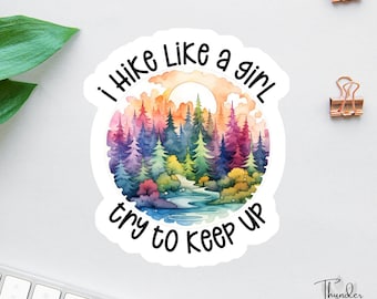 Puede incluir: Una pegatina blanca redonda con el texto "I Hike Like a Girl try to keep up". La pegatina presenta una ilustración de acuarela de un bosque con árboles coloridos, un río y un sol en el fondo.
