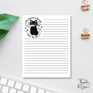 May include: A lined notepad with a black cat illustration holding a knife, surrounded by hearts. Text reads "The Horrors Persist But So Do I." The notepad is on a white surface with gold paper clips.