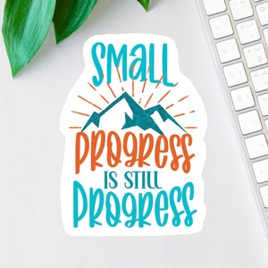 May include: A white sticker with a blue and orange mountain scene and the text "Small progress is still progress".