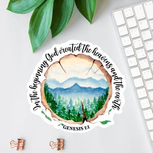 May include: A round sticker with a watercolor painting of a mountain landscape inside a tree ring. The sticker has the text "In the beginning God created the heavens and the earth" and "Genesis 1:1".