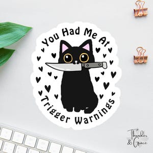 May include: A sticker featuring a black cat with yellow eyes holding a knife in its mouth. The sticker has the text "You Had Me At Trigger Warnings" in a circular design with black hearts.