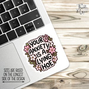 May include: A white sticker with the text "YOUR ANXIETY IS A LYING HOE" in a black, bold font, surrounded by pink flowers and green leaves. The sticker is on a laptop, with the text "SIZES ARE BASED ON THE LONGEST SIDE OF THE DESIGN".
