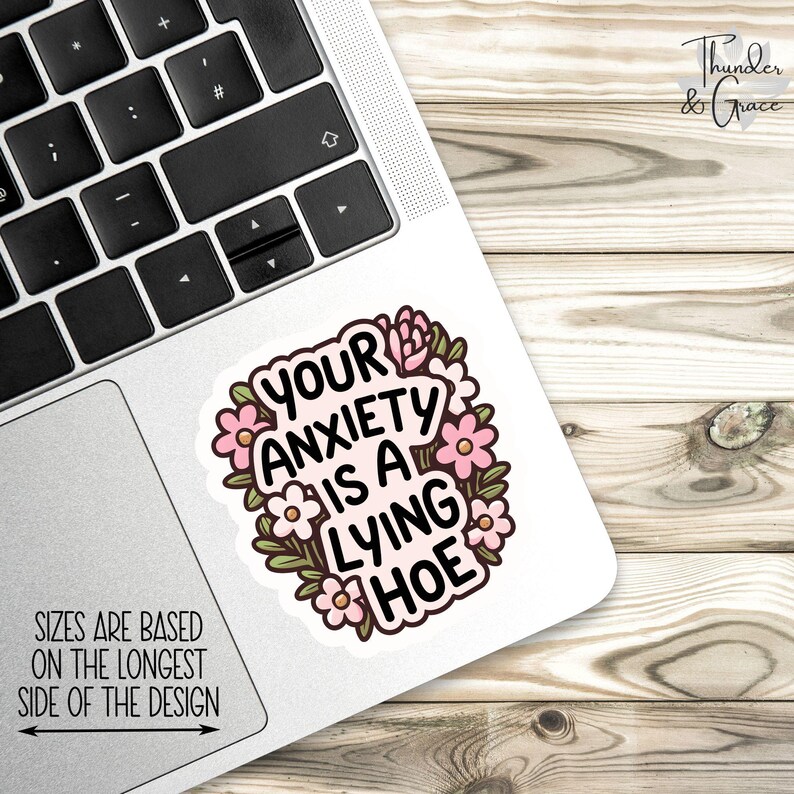 May include: A white sticker with the text "YOUR ANXIETY IS A LYING HOE" in a black, bold font, surrounded by pink flowers and green leaves. The sticker is on a laptop, with the text "SIZES ARE BASED ON THE LONGEST SIDE OF THE DESIGN".
