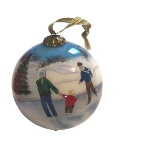 May include: A round Christmas ornament with a winter scene. The ornament features a family ice skating on a frozen pond, with a blue sky and a gold-colored string for hanging. The ornament is painted with a winter scene.