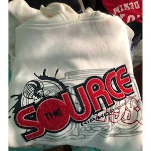 May include: White hoodie with a drawstring hood. The front features a large red and black embroidered design that reads "THE SOURCE" with "HIP HOP FOUNDATION" below. A graphic of a boombox is also visible.