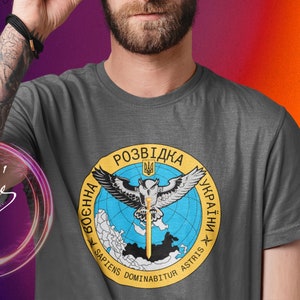 May include: A grey t-shirt with a yellow and blue circular graphic featuring an owl, a sword, and the text "Розвідка України" in Ukrainian. The text "Sapiens Dominabitur Astris" is also included in the graphic.