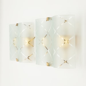 May include: Pair of frosted glass wall sconces with a geometric pattern. The sconces have a brass finish and are illuminated by light bulbs.