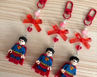 May include: Three identical keychains featuring a Superman minifigure with a blue and red suit and a red cape. Each keychain includes a red plastic lobster clasp, a translucent red bow, a small pink bead, and a small red heart-shaped charm.