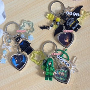 May include: Three metal keychains featuring superhero minifigures, heart-shaped photo charms, and various beads. One keychain includes a Nightwing figure, one features Batman with BATS letter beads, and one has Green Arrow with ARROW letter beads.