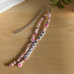 May include: A beaded bookmark with a silver metal top. The beads are white, pink, and gray, spelling out "LITTLE FOX" with heart accents. The bookmark is on a wooden surface.