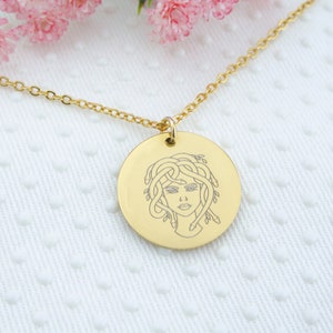May include: A gold necklace with a round pendant featuring a line drawing of Medusa, a Greek mythological figure with snakes for hair.