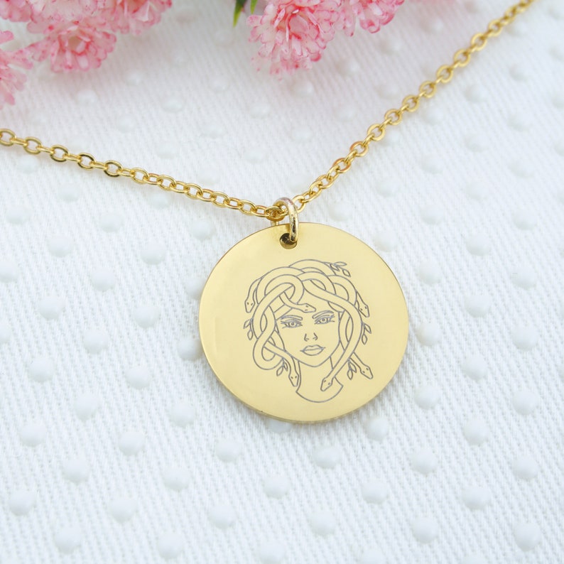 May include: A gold necklace with a round pendant featuring a line drawing of Medusa, a Greek mythological figure with snakes for hair.