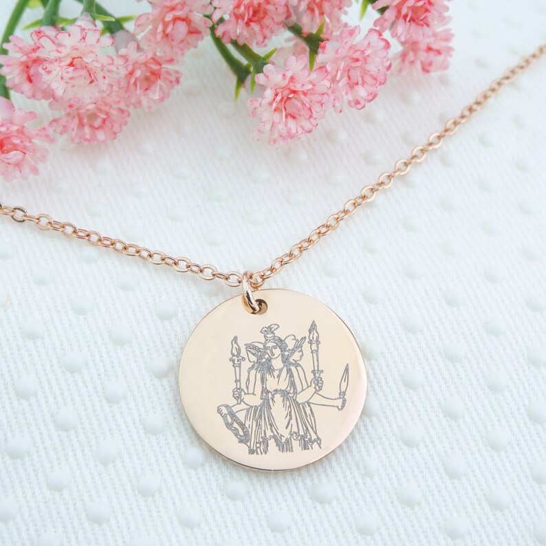 May include: A rose gold necklace with a round pendant featuring an engraved image of a three-headed goddess holding torches.