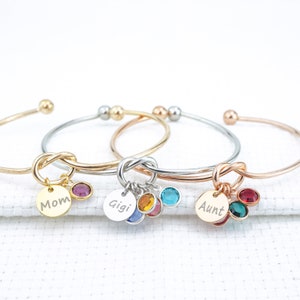 May include: Three gold, silver, and rose gold bangle bracelets with a knot design and a charm with a personalized engraved disc and colorful crystals. The charms read "Mom", "Gigi", and "Aunt".
