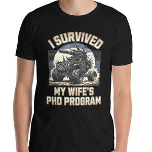 Puede incluir: Camiseta negra con el texto "I SURVIVED MY WIFE'S PHD PROGRAM" en blanco y amarillo. El gráfico presenta un vehículo de estilo militar con ruedas grandes y armas montadas, sobre un fondo de puesta de sol.
