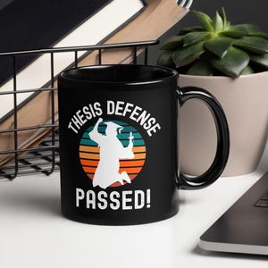 Peut inclure: Mug à café noir avec un graphique blanc d'une personne en toge et chapeau de graduation sautant en l'air avec le texte "Thesis Defense Passed!" dans un design de coucher de soleil rétro.
