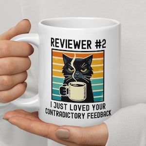 Może przedstawiać: Biały kubek ceramiczny z ilustracją czarnego kota trzymającego filiżankę kawy. Na kubku znajduje się napis "Reviewer #2 I just loved your contradictory feedback".
