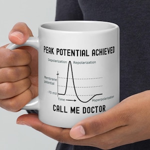 May include: White ceramic mug featuring the text "PEAK POTENTIAL ACHIEVED" and "CALL ME DOCTOR" in black. The mug also displays a diagram of a neuron's action potential. The mug is held by a person.