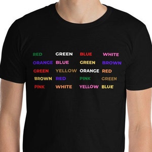 Può includere: Una maglietta nera con le parole "ROSSO", "VERDE", "BLU", "BIANCO", "ARANCIONE", "GIALLO", "ROSA" e "MARRONE" stampate in diversi colori. Le parole sono disposte in una griglia.