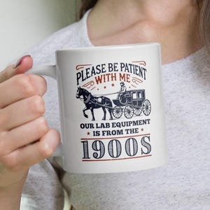 May include: White ceramic mug with the text "PLEASE BE PATIENT WITH ME" and "OUR LAB EQUIPMENT IS FROM THE 1900S" in red and black. The mug features a black and white illustration of a horse-drawn carriage.