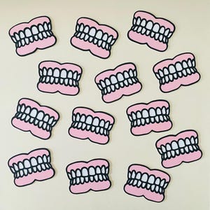 May include: A collection of cartoon teeth stickers. Each sticker features a set of white teeth outlined in black, set against a pink gum line. The stickers are scattered across a light beige background.